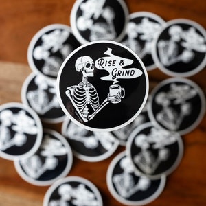 Sticker of a skeleton holding a coffee mug and in the steam coming off the coffee is text reading Rise & Grind