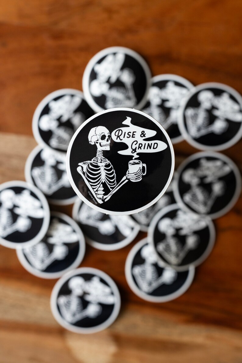Sticker of a skeleton holding a coffee mug and in the steam coming off the coffee is text reading Rise & Grind