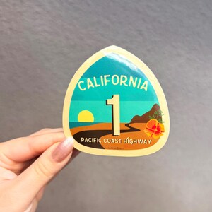 May include: A yellow and blue California Highway 1 sticker with a sunset and a poppy flower. The sticker says "California 1 Pacific Coast Highway".