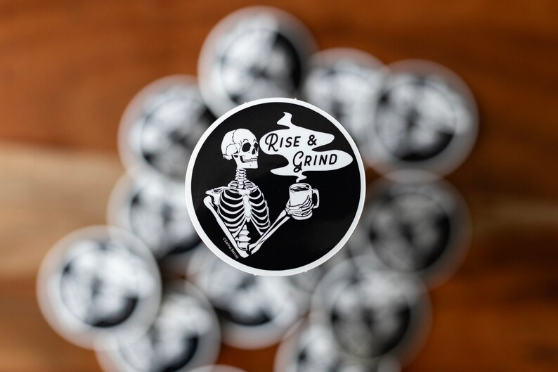 Sticker of a skeleton holding a coffee mug and in the steam coming off the coffee is text reading Rise & Grind