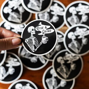 Sticker of a skeleton holding a coffee mug and in the steam coming off the coffee is text reading Rise & Grind