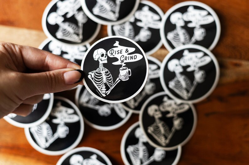 Sticker of a skeleton holding a coffee mug and in the steam coming off the coffee is text reading Rise & Grind