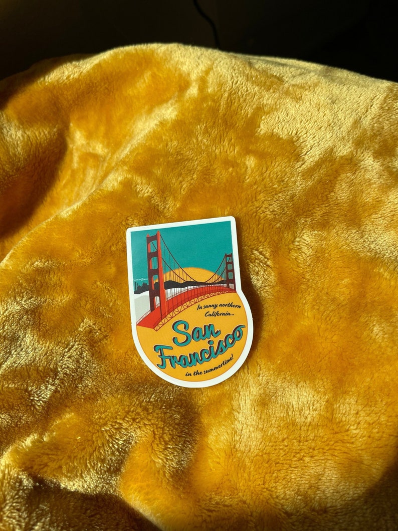 Sticker with a teal sky and yellow sun with the red Golden Gate Bridge. In teal script lettering it reads, San Francisco, and around it reads, In sunny northern California... in the summertime! Sticker rests on a yellow blanket.