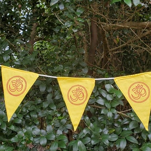 May include: A yellow bunting with red Om symbols on each flag. The bunting is hanging in a garden setting.