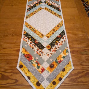 May include: A quilted table runner with a diamond pattern. The runner features various fabrics with sunflowers, fall leaves, and floral designs in yellow, orange, gray, and green. The edges are finished with a white border.