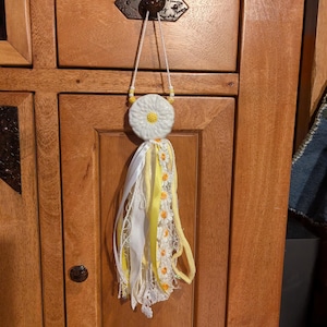May include: A decorative hanging ornament featuring a white ceramic daisy flower with a yellow center, suspended by a white cord with yellow beads. Below the flower hangs a cluster of white and yellow ribbons, lace, and small daisy trim.