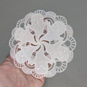 May include: A white, circular doily with a floral design. The doily is made of delicate lace and features a repeating pattern of stylized ghost-like shapes. The edges are scalloped, adding a decorative touch.