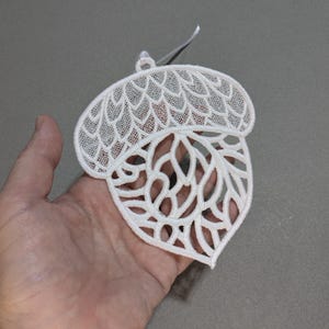 May include: White, openwork acorn ornament. The acorn is held in a hand and has a detailed, lacy design. The top of the acorn has a textured pattern, while the bottom has an open, leaf-like design. A small, white ribbon is attached for hanging.