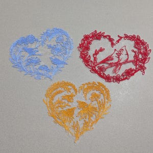 May include: Three embroidered heart-shaped patches. One is blue with birds and flowers, another is red with two birds on a branch, and the third is gold with birds and foliage. All are on a gray background.