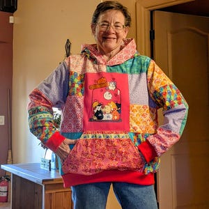 May include: A patchwork hoodie with a large front pocket and a hood. The hoodie features a vibrant mix of patterns and colors, including pink, blue, green, and orange. The front of the hoodie has a pink square with a jar of cats illustration.