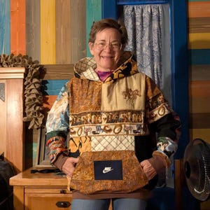 May include: A patchwork hoodie with a large front pocket featuring a Nike logo. The hoodie is made of various fabrics with horse and equestrian-themed patterns in shades of brown, beige, and blue. The sleeves are black and patterned.