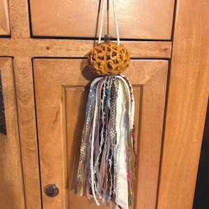 May include: A decorative hanging ornament featuring a carved, orange, pumpkin-shaped bead with an intricate openwork design. A long, multi-textured tassel made of various ribbons, yarns, and metallic threads in shades of cream, brown, and gold hangs below.