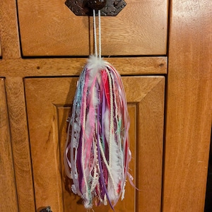 May include: A decorative hanging ornament composed of numerous vertical ribbons, fabric strips, and white feathers in shades of pink, purple, red, and white. The strands are gathered at the top and suspended by a white cord from a dark metal knob.