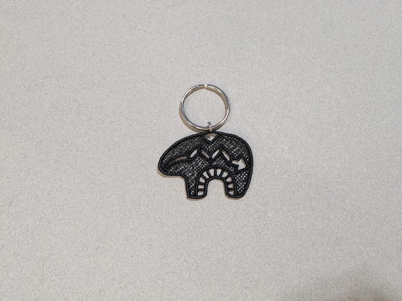 May include: Black metal keychain featuring a stylized bear design with intricate details. The bear is attached to a silver key ring.