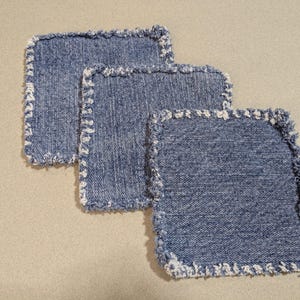 May include: Three square denim coasters with frayed edges. The coasters are made from blue denim fabric and have a light-colored, fringed border. They are arranged in a slightly overlapping pattern, showcasing their texture and design.
