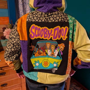 May include: A patchwork hoodie featuring a Scooby-Doo! graphic on the back. The jacket has a black base with the cartoon characters in the Mystery Machine. The hood and sleeves are various colors and patterns, including purple, orange, and green.