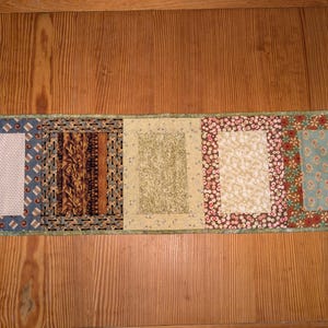 May include: A patchwork table runner with five different fabric panels. The panels feature various patterns in shades of blue, brown, green, and beige. The runner is approximately 15 inches wide and 40 inches long.
