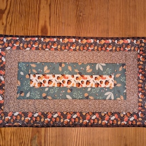 May include: Rectangular quilted table runner with an autumn theme. The design features a border of acorns and leaves, with a central panel of alternating patterns of acorns, leaves, and a speckled brown fabric. The color palette includes brown, orange, and green.