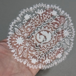 May include: A delicate, white lace doily with an intricate floral and leaf pattern. The center features a detailed design within an oval frame. The doily is held in a hand, showcasing its fine details and openwork design.