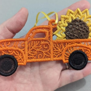 May include: An orange lace truck ornament featuring a detailed floral pattern, two solid black circular wheels, and a large yellow sunflower with a dark brown center in the truck bed, complete with a thin yellow ribbon loop for hanging.