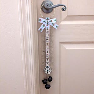 May include: A Christmas door hanger with a white ribbon bow featuring green Christmas trees and red berries. The word "CHRISTMAS" is spelled out vertically on a burlap strip, with a snowflake and three black bells at the bottom.