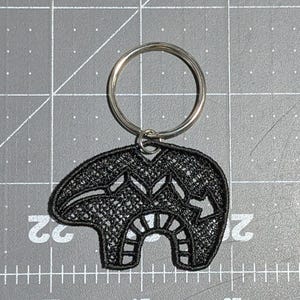 May include: Black embroidered bear keychain with a silver key ring. The bear is a traditional design with intricate details.