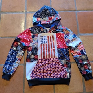 May include: A patriotic hoodie with a patchwork design featuring American flags, stars, and fireworks. The front showcases a sequined American flag. The sleeves and hood have a navy blue background with white stars and red, white, and blue patterns. The bottom and cuffs are black.