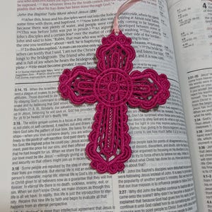 May include: A magenta-colored, openwork cross pendant with a delicate design, hanging over an open book. The cross is made of a woven material and is suspended by a pink ribbon.