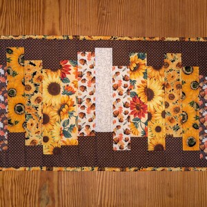 May include: Rectangular quilted table runner featuring a patchwork design with sunflowers, acorns, and mushroom patterns. The runner has a brown polka dot border and a central white stripe. The color palette includes yellow, orange, brown, and black.