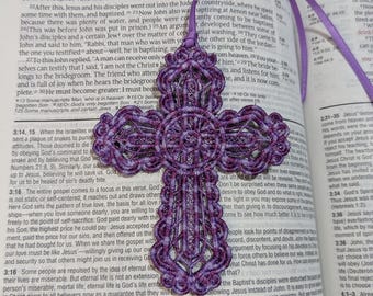 May include: A purple macrame cross with a decorative design, hanging on a purple ribbon. The cross is displayed over an open book with text. A religious item for faith-based decor.