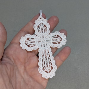 May include: A white, ornate lace cross with a scalloped edge and a small ribbon loop for hanging. The cross features intricate openwork patterns and a central circular design. The delicate details suggest a religious or decorative item.