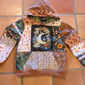 May include: A patchwork hoodie featuring a central rectangular panel with a crescent moon, floral, and mushroom graphic. The garment is constructed from various fabric squares in earthy tones, including orange, green, and brown, with a solid brown hem and cuffs.