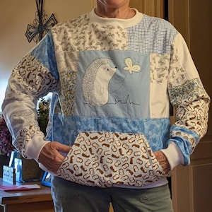 May include: A patchwork sweatshirt with a hedgehog and butterfly design. The sweatshirt features a white crew neck, white cuffs, and a kangaroo pocket. The sleeves and body are made of various patterned fabrics in blue, white, and brown.