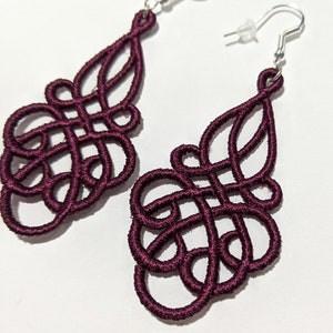 May include: A pair of burgundy dangle earrings with a delicate, intricate design. The earrings are made of a woven material and have silver hooks.
