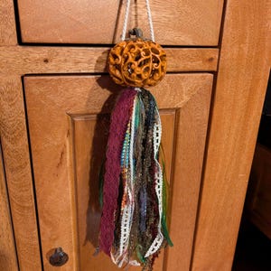 May include: A decorative hanging ornament featuring a carved orange pumpkin with intricate scrollwork patterns. A white cord suspends the pumpkin, which is adorned with a long, multi-textured tassel made of various fabric strips, lace, and ribbons.
