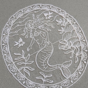 May include: White embroidered patch featuring a mermaid design within a circular frame. The mermaid is surrounded by aquatic plants and a fish. The border has a decorative floral pattern. The patch is on a gray background.