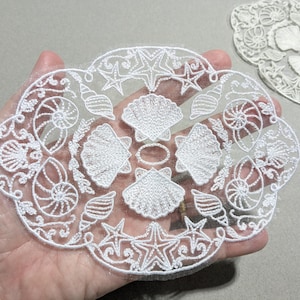 May include: A white lace appliqué featuring an intricate embroidered design of various seashells, starfish, and swirling patterns on a sheer mesh background. The oval-shaped piece displays detailed stitching of scallop shells and spiral conch shells.