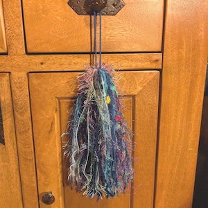 May include: A decorative tassel made of various textured yarns in shades of blue, purple, and pink, featuring metallic threads and small colorful accents. The tassel hangs from a blue cord attached to a dark metal knob on a wooden cabinet door.