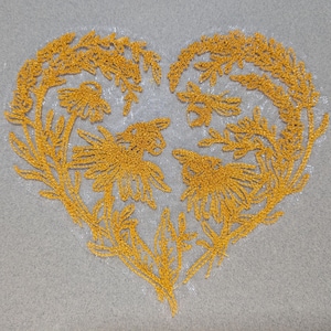 May include: A heart-shaped design embroidered with gold thread on a gray background. The embroidery features floral and botanical elements, creating a delicate and detailed pattern. The design is centered and fills the heart shape.