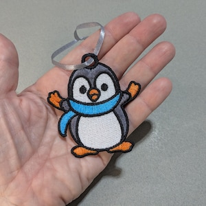 May include: Embroidered penguin decoration with a grey head, white body, and orange feet and beak. The penguin wears a blue scarf and has a silver ribbon for hanging. A charming, whimsical design.