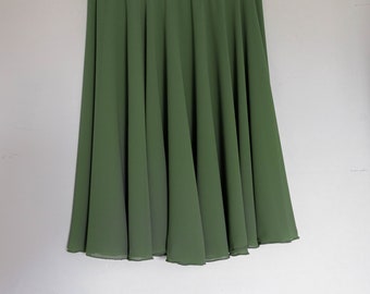 May include: A flowing, olive green skirt hangs from a wooden and metal hanger. The skirt has a flared, A-line silhouette, with soft folds and a slightly sheer appearance. The fabric appears lightweight and suitable for warmer weather.