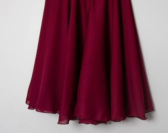 May include: A deep burgundy knee-length skirt with a flared, flowing silhouette and a gathered elastic waistband. The garment is made of a lightweight, semi-sheer fabric and is displayed hanging from a wooden and metal clothes hanger.