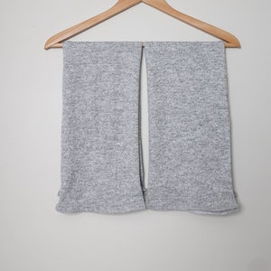 Op de afbeelding: Twee lichtgrijze sjaals hangen aan een houten kleerhanger tegen een witte muur. De sjaals hebben een textuur en zijn gemaakt van een zacht materiaal. De hanger is gemaakt van lichtgekleurd hout en heeft een metalen haak.