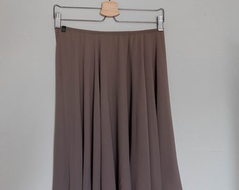 May include: A knee-length, taupe-colored skirt hangs from a wooden and metal hanger against a light gray wall. The skirt has a flowing, flared design, with soft folds and a slightly sheer appearance. The fabric drapes gracefully.