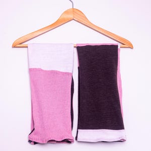 May include: A pair of ribbed knit leg warmers hanging on a wooden hanger. The left leg warmer features a light pink top section and a darker pink ribbed bottom. The right leg warmer is dark charcoal gray with a light pink band at the bottom edge.