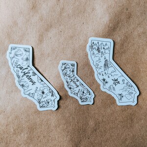 May include: Three white stickers in the shape of California. Two stickers feature floral designs and the word "California" in script. The third sticker depicts a landscape with mountains, trees, and buildings. All stickers have black line art.