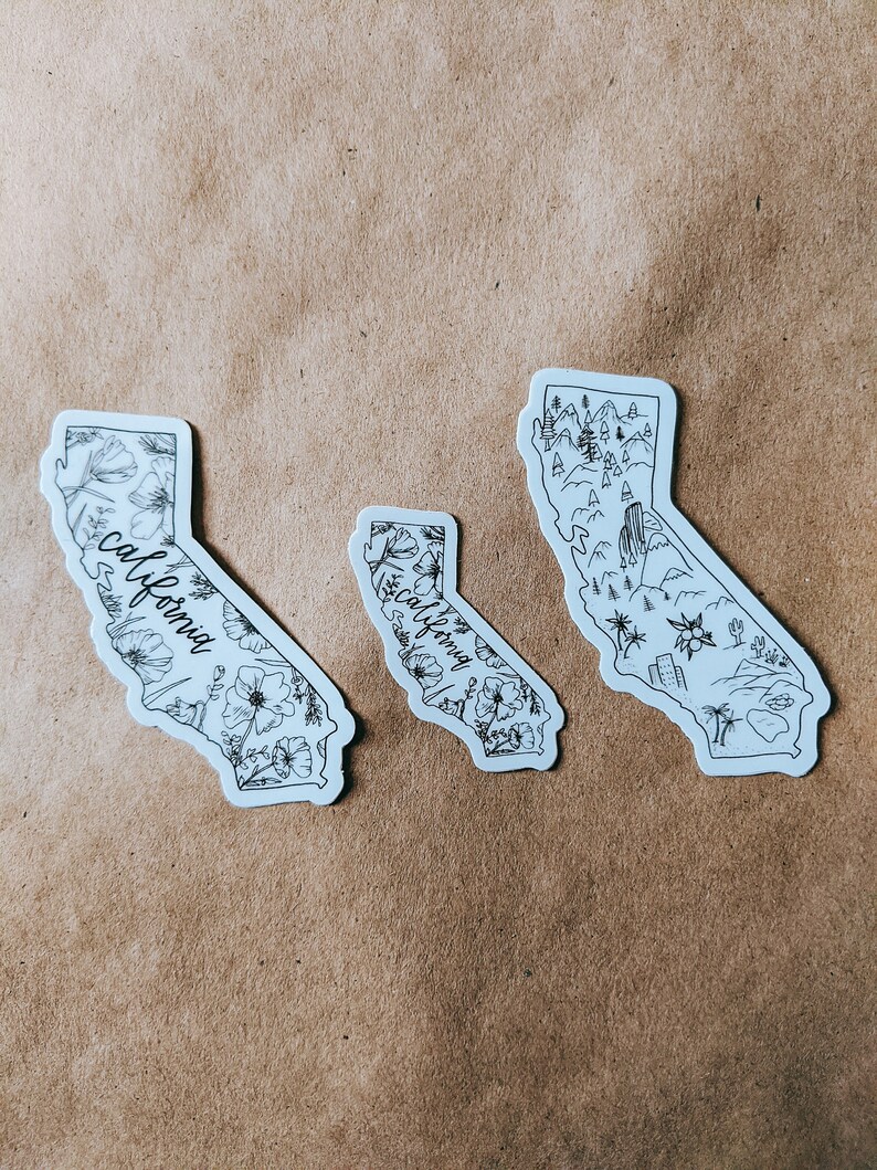 May include: Three white stickers in the shape of California. Two stickers feature floral designs and the word "California" in script. The third sticker depicts a landscape with mountains, trees, and buildings. All stickers have black line art.