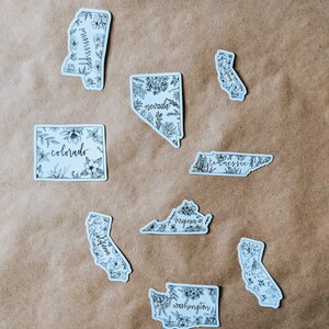 May include: A collection of white stickers featuring state outlines, each filled with floral illustrations and state names in a handwritten font. States include Mississippi, Nevada, Colorado, California, Tennessee, Virginia, and Washington.