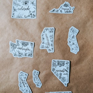 May include: A collection of white stickers featuring state outlines, each adorned with floral illustrations and state names in a handwritten font. States include Colorado, Virginia, Washington, Mississippi, California, Nevada, and Tennessee.