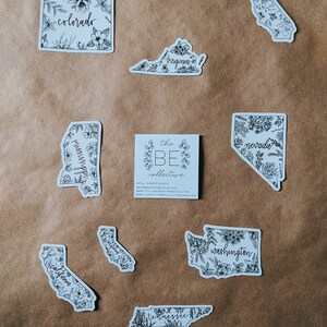 May include: A collection of white stickers featuring state outlines with floral illustrations and state names in cursive. States include Colorado, Virginia, Mississippi, Nevada, California, Tennessee, and Washington. A small card with the text "the BE collective" is also present.